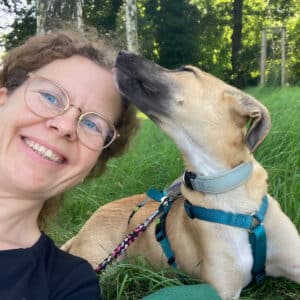Kornelia und Hund Lotte im Gras. Lotte schnüffelt an Kornelias Kopf.
